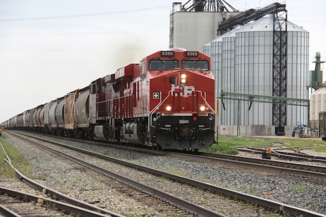 A freight train with a red locomotive is traveling on railway tracks beside large, industrial silos and structures. The sky is overcast, and the area appears to be part of an industrial zone.