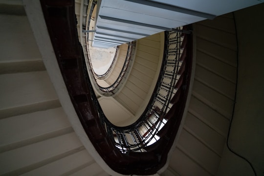 A winding spiral staircase viewed from an upward angle, highlighting concentric circles formed by the steps and railings. The structure features a mix of light and dark hues, creating an intriguing pattern.