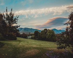 A serene landscape featuring a lush green lawn bordered by various trees and shrubs. In the background, a range of mountains can be seen under a partly cloudy sky. The clouds are tinged with soft hues from the setting or rising sun.