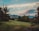 A serene landscape featuring a lush green lawn bordered by various trees and shrubs. In the background, a range of mountains can be seen under a partly cloudy sky. The clouds are tinged with soft hues from the setting or rising sun.