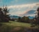 A serene landscape featuring a lush green lawn bordered by various trees and shrubs. In the background, a range of mountains can be seen under a partly cloudy sky. The clouds are tinged with soft hues from the setting or rising sun.