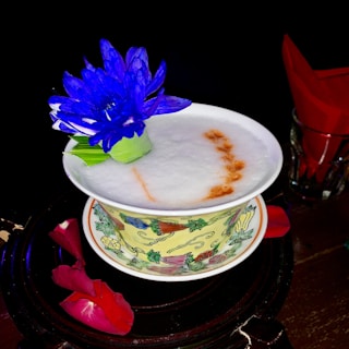 A decorative cup filled with a foamy beverage, adorned with a bright blue flower. The cup features intricate floral patterns and rests on a saucer, accompanied by scattered red rose petals and a glass holding red napkins.