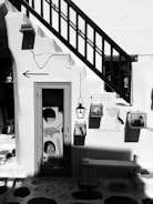 A black and white photograph of a quaint storefront with a staircase passing over a door. The door has artistic decorations including animal and abstract designs. There are signs, one saying 'THIS IS NOT A GALLERY' with bird illustrations nearby. The setting feels artistic with an eclectic and creative atmosphere.