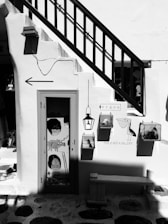 A black and white photograph of a quaint storefront with a staircase passing over a door. The door has artistic decorations including animal and abstract designs. There are signs, one saying 'THIS IS NOT A GALLERY' with bird illustrations nearby. The setting feels artistic with an eclectic and creative atmosphere.