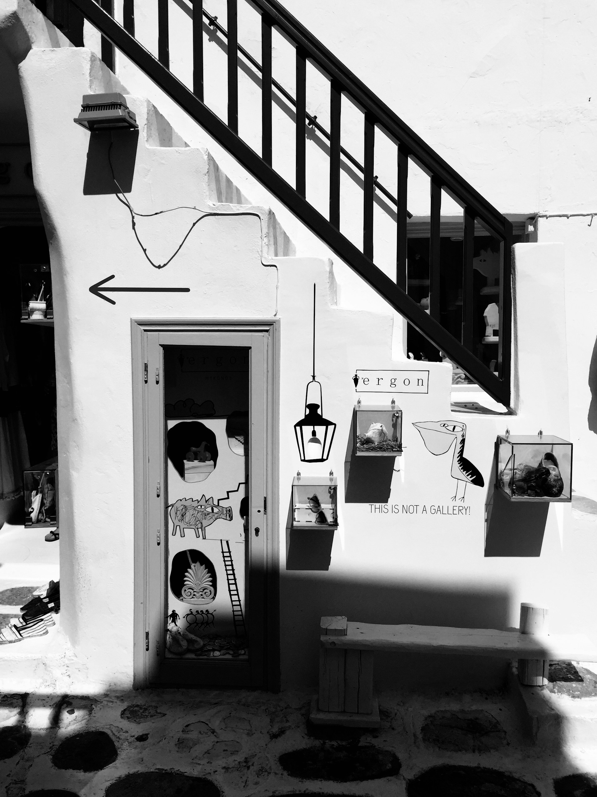 A black and white photograph of a quaint storefront with a staircase passing over a door. The door has artistic decorations including animal and abstract designs. There are signs, one saying 'THIS IS NOT A GALLERY' with bird illustrations nearby. The setting feels artistic with an eclectic and creative atmosphere.