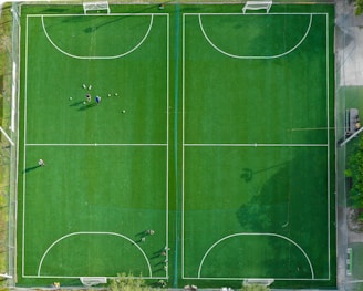 An aerial view of two adjacent soccer fields with neatly marked pitch lines. The fields are surrounded by a netting border. Several people are scattered across the left field, some standing near a group of soccer balls, indicating a practice session or a casual game.