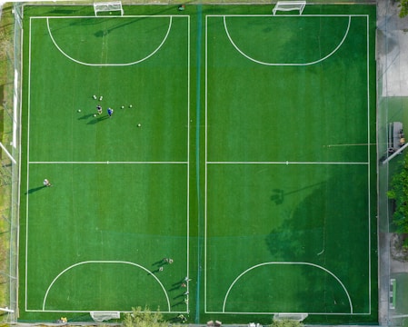 An aerial view of two adjacent soccer fields with neatly marked pitch lines. The fields are surrounded by a netting border. Several people are scattered across the left field, some standing near a group of soccer balls, indicating a practice session or a casual game.