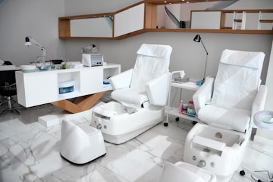 The image depicts a modern and clean spa or salon environment. There are two comfortable, white pedicure chairs with matching foot baths. The floor is covered with white marble tiles. In the background, a shelving unit holds various spa equipment and containers, and there's a mirror with wooden accents above. The space is well-lit with several desk lamps placed strategically around.