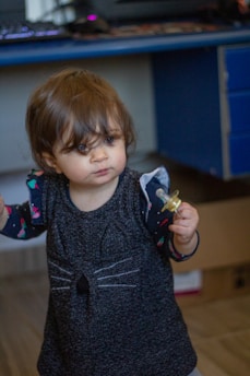 A young child holding a pacifier stands indoors. The child is wearing a dress with a cat design on it, featuring whiskers on the chest. The background includes a blue desk with a computer and some out-of-focus objects.