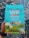 A colorful coffee packaging held by a person, featuring vibrant illustrations of a bird, mountains, and small houses. The text highlights that it is Trader Joe's El Salvador Santa Ana Small Lot Coffee, with 100% Arabica whole bean, medium roast, and notes of dark chocolate, apple, and butterscotch. The packaging is predominantly turquoise with accents of red, yellow, and green.