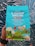 A colorful coffee packaging held by a person, featuring vibrant illustrations of a bird, mountains, and small houses. The text highlights that it is Trader Joe's El Salvador Santa Ana Small Lot Coffee, with 100% Arabica whole bean, medium roast, and notes of dark chocolate, apple, and butterscotch. The packaging is predominantly turquoise with accents of red, yellow, and green.