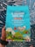 A colorful coffee packaging held by a person, featuring vibrant illustrations of a bird, mountains, and small houses. The text highlights that it is Trader Joe's El Salvador Santa Ana Small Lot Coffee, with 100% Arabica whole bean, medium roast, and notes of dark chocolate, apple, and butterscotch. The packaging is predominantly turquoise with accents of red, yellow, and green.