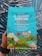 A colorful coffee packaging held by a person, featuring vibrant illustrations of a bird, mountains, and small houses. The text highlights that it is Trader Joe's El Salvador Santa Ana Small Lot Coffee, with 100% Arabica whole bean, medium roast, and notes of dark chocolate, apple, and butterscotch. The packaging is predominantly turquoise with accents of red, yellow, and green.