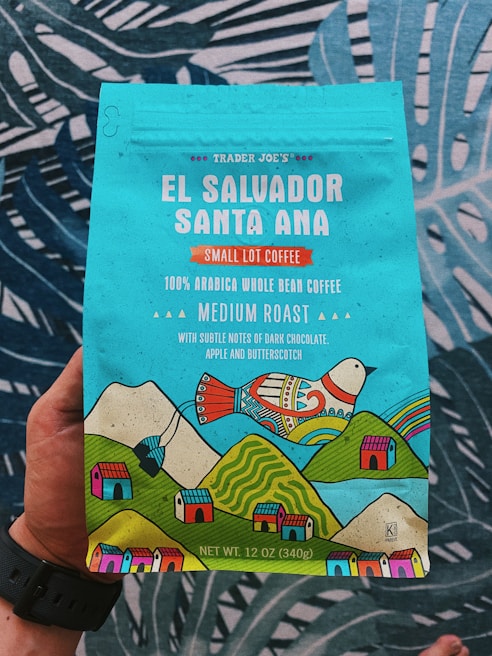 A colorful coffee packaging held by a person, featuring vibrant illustrations of a bird, mountains, and small houses. The text highlights that it is Trader Joe's El Salvador Santa Ana Small Lot Coffee, with 100% Arabica whole bean, medium roast, and notes of dark chocolate, apple, and butterscotch. The packaging is predominantly turquoise with accents of red, yellow, and green.