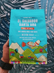 A colorful coffee packaging held by a person, featuring vibrant illustrations of a bird, mountains, and small houses. The text highlights that it is Trader Joe's El Salvador Santa Ana Small Lot Coffee, with 100% Arabica whole bean, medium roast, and notes of dark chocolate, apple, and butterscotch. The packaging is predominantly turquoise with accents of red, yellow, and green.