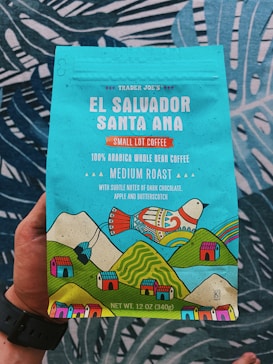 A colorful coffee packaging held by a person, featuring vibrant illustrations of a bird, mountains, and small houses. The text highlights that it is Trader Joe's El Salvador Santa Ana Small Lot Coffee, with 100% Arabica whole bean, medium roast, and notes of dark chocolate, apple, and butterscotch. The packaging is predominantly turquoise with accents of red, yellow, and green.