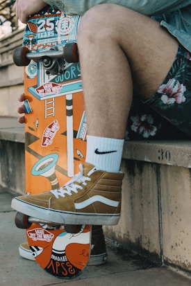A person is sitting on a concrete ledge, resting a skateboard vertically against it. The skateboard has a colorful graphic design with various stickers and an orange base. The person is wearing white socks and brown high-top sneakers.