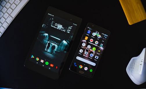 A tablet and smartphone are placed on a dark surface along with parts of a keyboard and a white computer mouse. The tablet displays an urban night scene with two silhouetted figures, while the smartphone shows various colorful app icons on its screen.