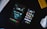 A tablet and smartphone are placed on a dark surface along with parts of a keyboard and a white computer mouse. The tablet displays an urban night scene with two silhouetted figures, while the smartphone shows various colorful app icons on its screen.