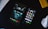 A tablet and smartphone are placed on a dark surface along with parts of a keyboard and a white computer mouse. The tablet displays an urban night scene with two silhouetted figures, while the smartphone shows various colorful app icons on its screen.