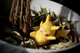 A small, yellow Pikachu figurine is nestled among dry leaves in a white plant pot. The figurine has closed eyes and a relaxed expression, positioned beside the base of a small tree. Natural lighting casts shadows, adding depth to the scene.