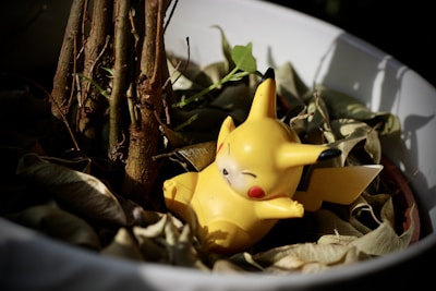 A small, yellow Pikachu figurine is nestled among dry leaves in a white plant pot. The figurine has closed eyes and a relaxed expression, positioned beside the base of a small tree. Natural lighting casts shadows, adding depth to the scene.