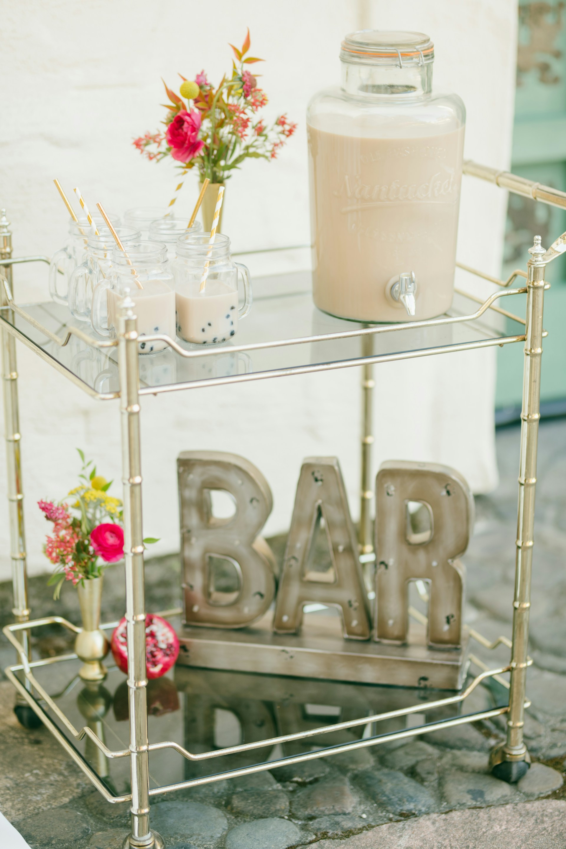 A bar cart with a large glass dispenser filled with a creamy beverage, likely milk tea, on the top shelf. Beside it are four glass jars each with a straw, containing a similar drink. Pink and red flowers in small vases add color to the scene. The lower level of the cart holds a decorative metallic 'BAR' sign and a pomegranate near the flowers.