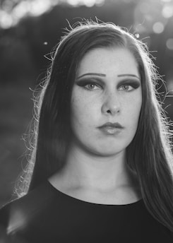 A monochromatic portrait of a young person with straight long hair, emphasizing dramatic eye makeup and a calm expression. The soft background bokeh creates a serene and introspective atmosphere.