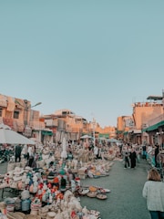 A bustling outdoor market scene with numerous stalls selling a variety of woven baskets, pottery, and textiles. The market is crowded with people walking and browsing the items. Sunlit buildings with colorful architecture line the background, and the sky is clear and blue.