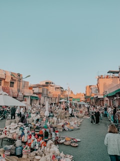A bustling outdoor market scene with numerous stalls selling a variety of woven baskets, pottery, and textiles. The market is crowded with people walking and browsing the items. Sunlit buildings with colorful architecture line the background, and the sky is clear and blue.