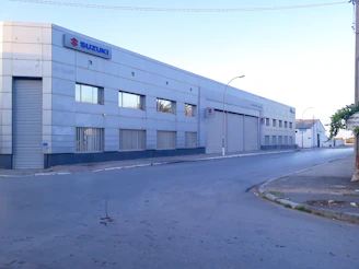 A large, industrial-style building with a metal facade and several small windows. The building has a prominent Suzuki logo on the upper corner. There is an empty street in front of it, and the area appears quiet with no visible people or vehicles.