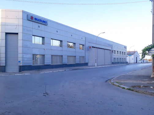 A large, industrial-style building with a metal facade and several small windows. The building has a prominent Suzuki logo on the upper corner. There is an empty street in front of it, and the area appears quiet with no visible people or vehicles.