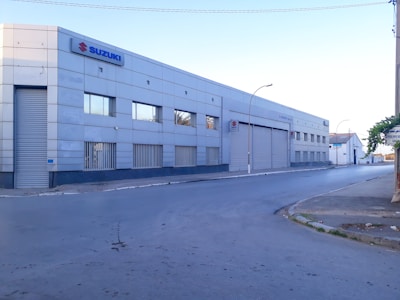 A large, industrial-style building with a metal facade and several small windows. The building has a prominent Suzuki logo on the upper corner. There is an empty street in front of it, and the area appears quiet with no visible people or vehicles.