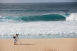 A photographer with a backpack is standing on a sandy beach using a tripod-mounted camera to capture the scene. In the distance, a large ocean wave is approaching the shore, and a solitary person is visible in the water.