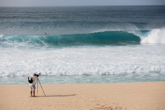 A photographer with a backpack is standing on a sandy beach using a tripod-mounted camera to capture the scene. In the distance, a large ocean wave is approaching the shore, and a solitary person is visible in the water.
