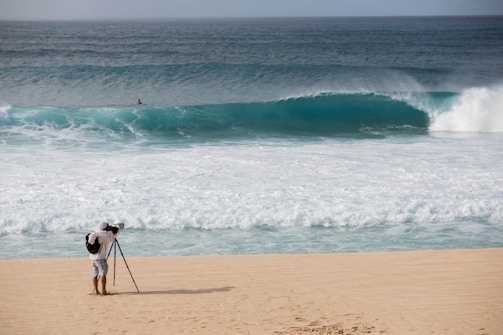 A photographer with a backpack is standing on a sandy beach using a tripod-mounted camera to capture the scene. In the distance, a large ocean wave is approaching the shore, and a solitary person is visible in the water.