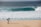 A photographer with a backpack is standing on a sandy beach using a tripod-mounted camera to capture the scene. In the distance, a large ocean wave is approaching the shore, and a solitary person is visible in the water.