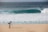 A photographer with a backpack is standing on a sandy beach using a tripod-mounted camera to capture the scene. In the distance, a large ocean wave is approaching the shore, and a solitary person is visible in the water.