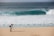 A photographer with a backpack is standing on a sandy beach using a tripod-mounted camera to capture the scene. In the distance, a large ocean wave is approaching the shore, and a solitary person is visible in the water.