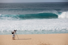 A photographer with a backpack is standing on a sandy beach using a tripod-mounted camera to capture the scene. In the distance, a large ocean wave is approaching the shore, and a solitary person is visible in the water.