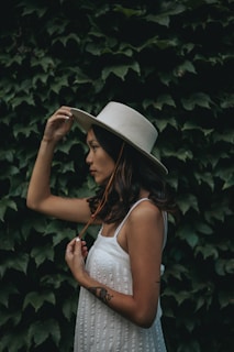 A woman in profile wearing a white hat and a white dress stands in front of a dense green ivy wall. She has long hair and holds the brim of her hat with one hand while her other hand gently touches the string of the hat. There is a tattoo visible on her forearm.