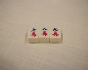 Three Mahjong tiles are placed in a row on a textured, off-white surface. The tiles each display Chinese characters in black and red ink.