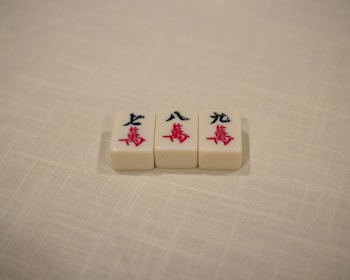 Three Mahjong tiles are placed in a row on a textured, off-white surface. The tiles each display Chinese characters in black and red ink.
