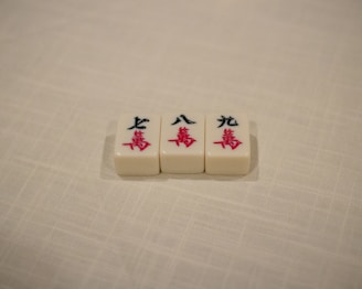 Three Mahjong tiles are placed in a row on a textured, off-white surface. The tiles each display Chinese characters in black and red ink.