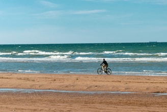 A biker riding along a sandy beach with waves crashing in the background.