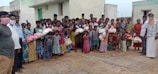 A large group of people, including men, women, and children, stands together outside in front of concrete buildings. Many are holding bags, possibly containing supplies or food. Some individuals are wearing face coverings.