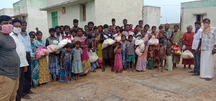 A large group of people, including men, women, and children, stands together outside in front of concrete buildings. Many are holding bags, possibly containing supplies or food. Some individuals are wearing face coverings.