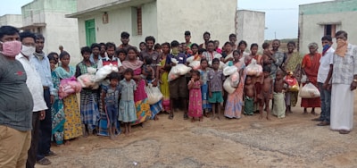 A large group of people, including men, women, and children, stands together outside in front of concrete buildings. Many are holding bags, possibly containing supplies or food. Some individuals are wearing face coverings.