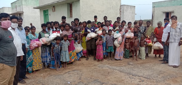 A large group of people, including men, women, and children, stands together outside in front of concrete buildings. Many are holding bags, possibly containing supplies or food. Some individuals are wearing face coverings.