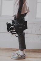 A person is holding a professional video camera with both hands, wearing rolled-up pants, colorful sneakers, and white socks. The background consists of a white brick wall and wooden floor.